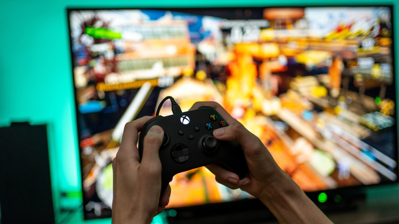 Hands holding an Xbox Series X controller with a TV displaying a videogame in the background.