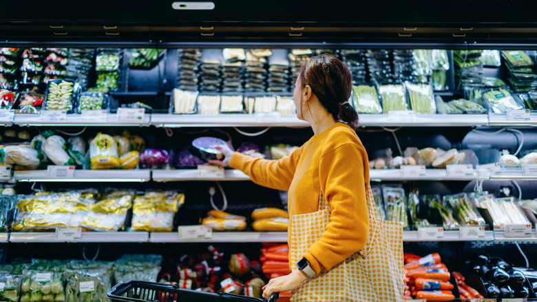 Young Asian woman with shopping cart, carrying a reusable shopping bag, shopping for fresh organic fruits and vegetables in supermarket.