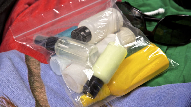 A messy ziplock bag filled with various small toiletry bottles sitting on a pile of clothing next to a pair of sunglasses