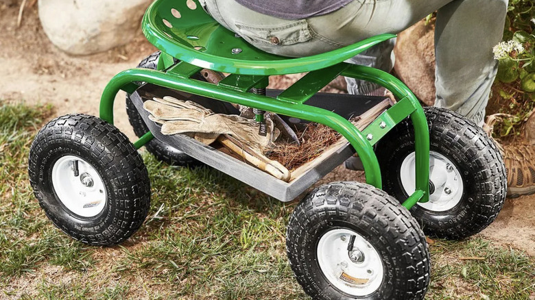 A person sitting on a the rolling work seat from Harbor Freight next to a garden, with gloves and tools in the bin below the seat