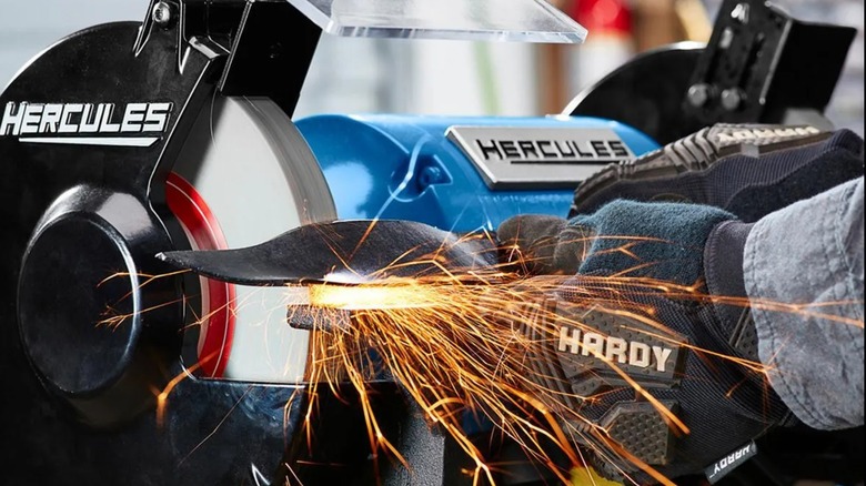A worker, wearing safety gloves, sharpening a lawn mower blade with a Hercules bench grinder.