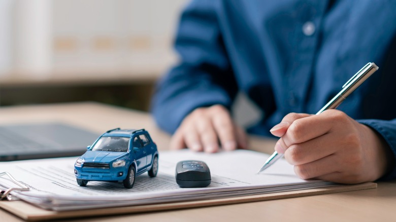 A person signing paperwork with a model car and car key fob placed on the paper.