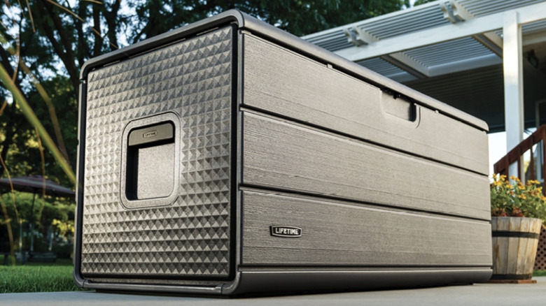 The Lifetime 170-Gallon Resin Outdoor Storage Deck Box on a patio with a pot of flowers next to it