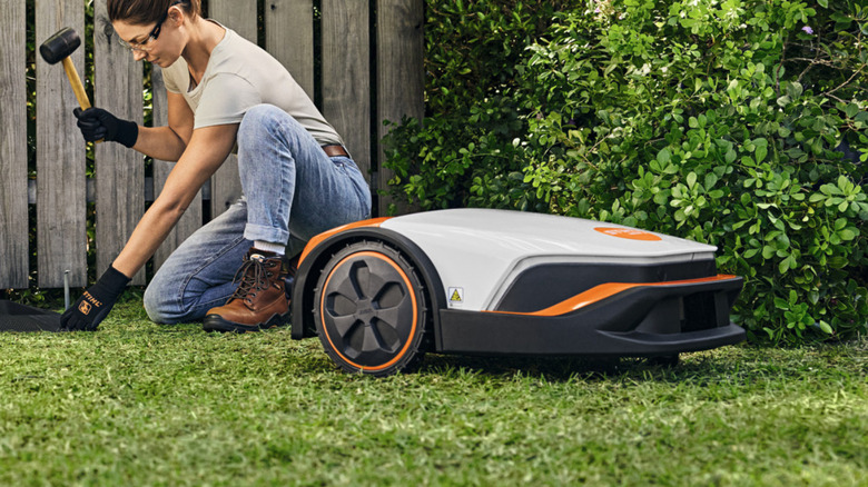 An android pushing a blue lawn mower across a green lawn in front of a white house surrounded by trees.