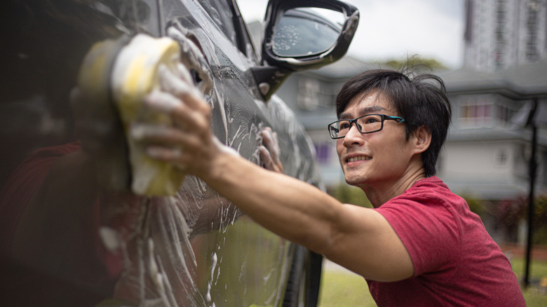 A smiling person using a yellow sponge to wash a gray vehicle