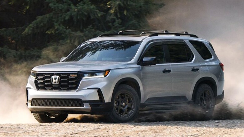 A 2026 silver Honda Pilot SUV kicks up a trail of dust while driving through a forested dirt road, capturing its off-road capability.