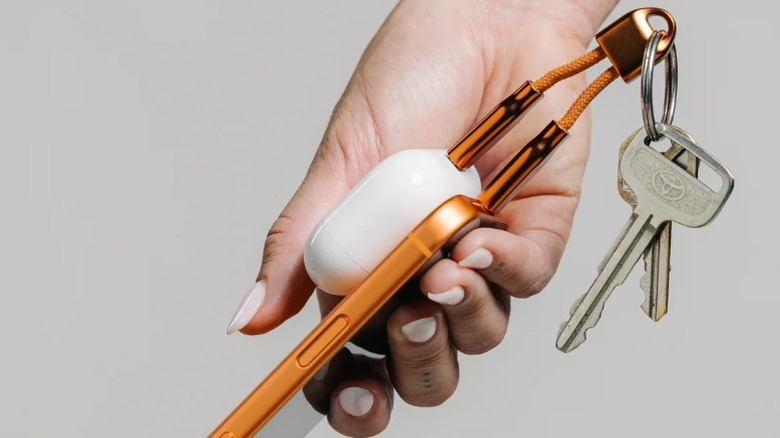 Hand holding orange smartphone and earbuds case, connected via an orange Nomad ChargeKey cable