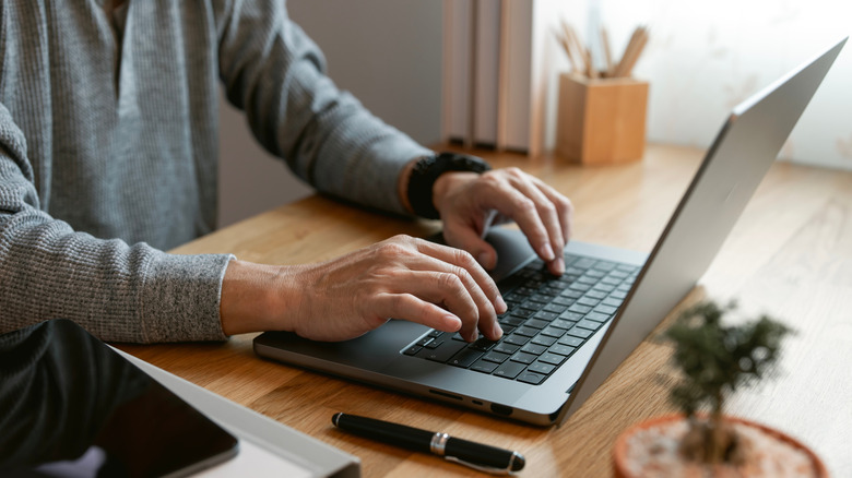 A close-up of a person using a larger MacBook on a wooden desk, pen and iPad to their right