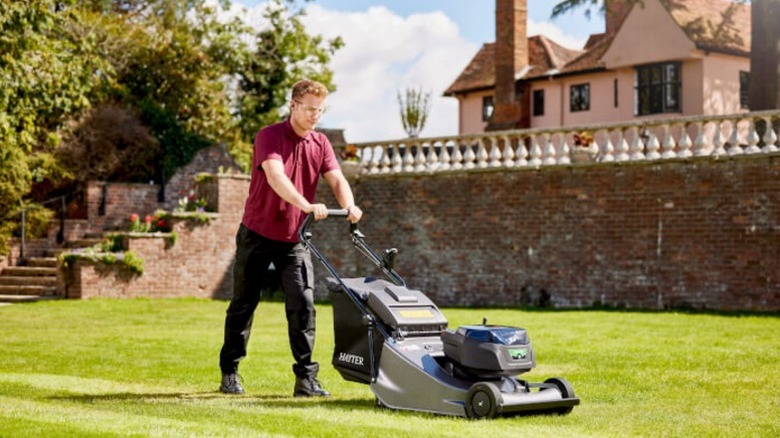 Person mowing a lawn with a Hayter lawn mower, a brick wall and English country house in the background