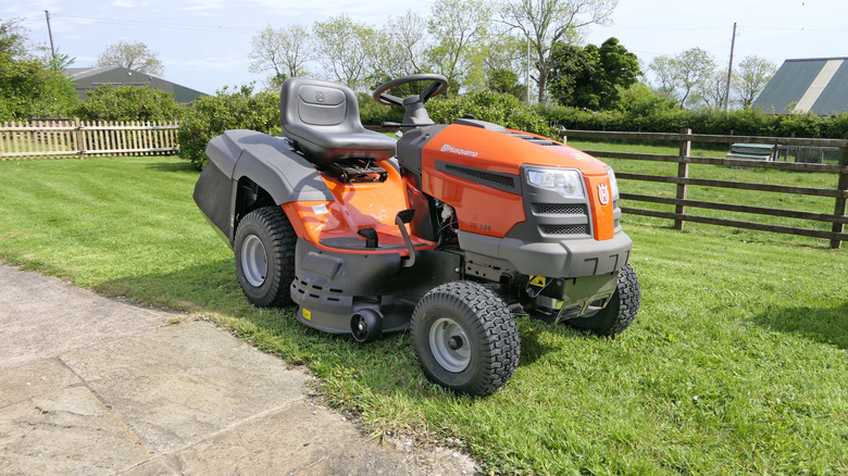 A Husqvarna lawn tractor parked on a lawn next between a wooden fence and a concrete driveway, front 3/4 view