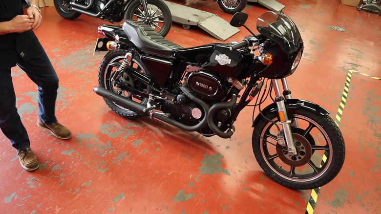 A black Harley Davidson XCLR cafe racer motorbike from the late 1970s standing on an orange floor with a man next to it.