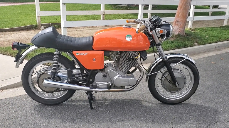 A 1972 Laverda 750 SF motorcycle in cafe racer styling finished in a bright orange color parked on a country road with a white fence in the back.