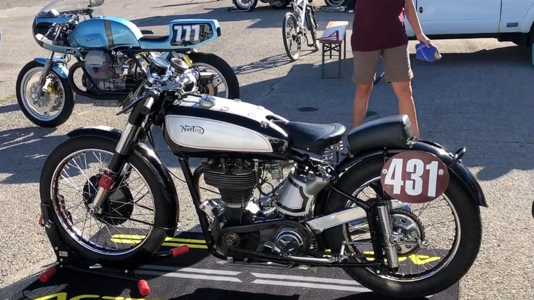 A 1947 Norton Manx 500 finished in white standing on tarmac, with a blue cafe racer bike behind it.
