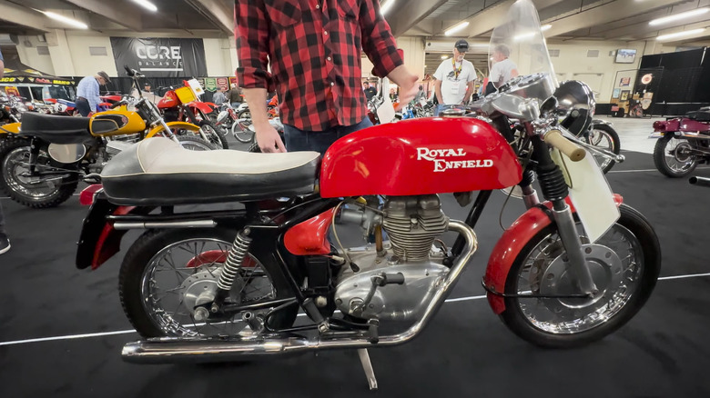 A red and chrome silver 1966 Royal Enfield Continental GT 250 cafe racer on an indoor auction floor with other bikes in the backdrop.