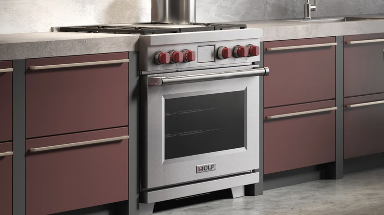 A Wolf range with red knobs set in modern dark red cabinets with a light countertop and backsplash