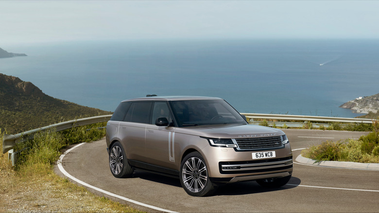 Front 3/4 view of a gold Range Rover on a mountain road, sea in the background