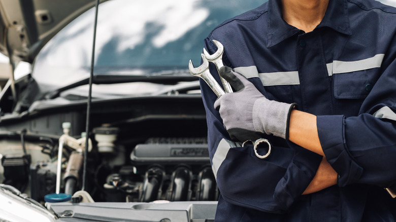 Car mechanic holding two wrenches and a car with hood open in background