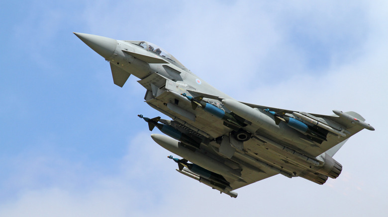 Eurofighter Typhoon fighter jet seen from below left displaying multiple hard points loaded with missiles and external fuel tanks