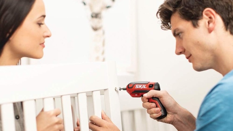 A woman holds some flat-pack furniture in place while a man uses a Skil mini-screwdriver to screw it together.