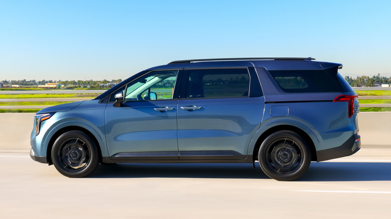 Side view of blue 2027 Kia Carnival driving on highway.