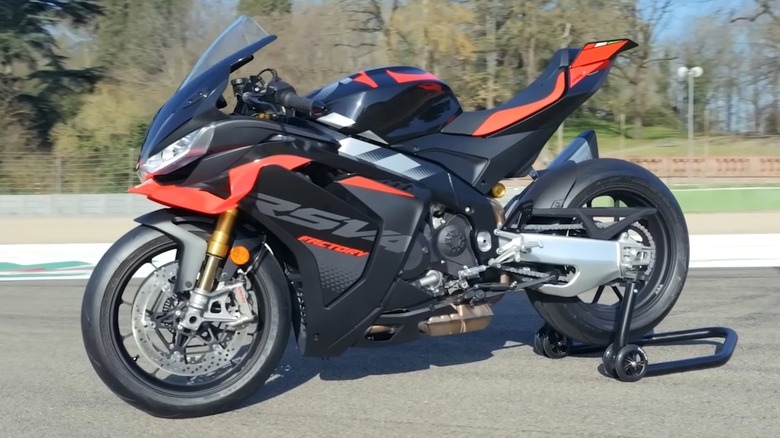 A close-up side view of an Aprilia RSV4 Factory sportbike parked on a race track, showing its aerodynamic design and red accents.