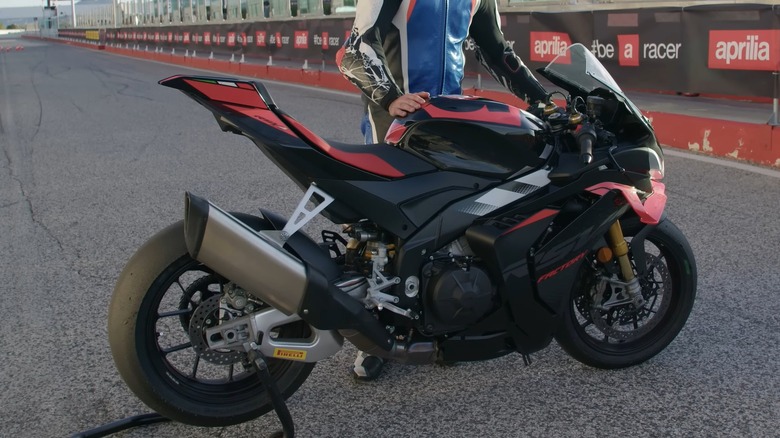 A rider in a blue and red racing suit standing beside an Aprilia RSV4 at a racetrack.