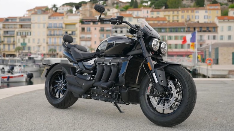 A 2025 black Triumph Rocket 3 parked near a harbor with European-style buildings in the background.