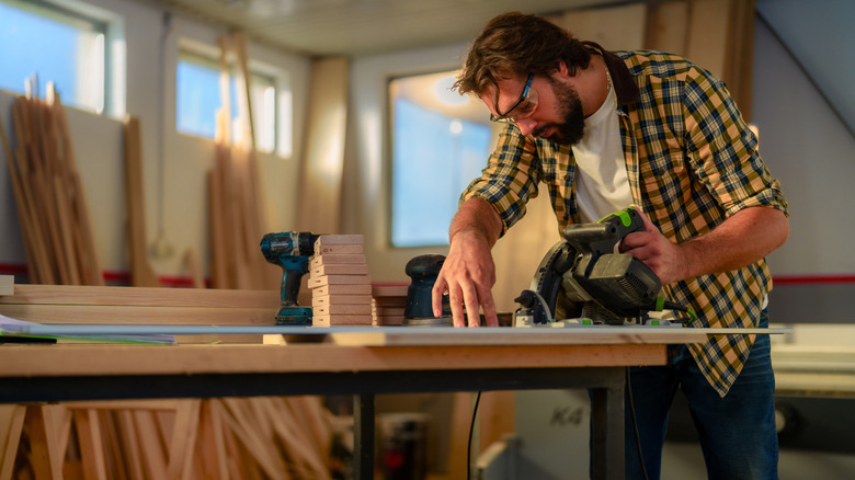 A man in a shop uses a circular saw while surrounded by wooden planks