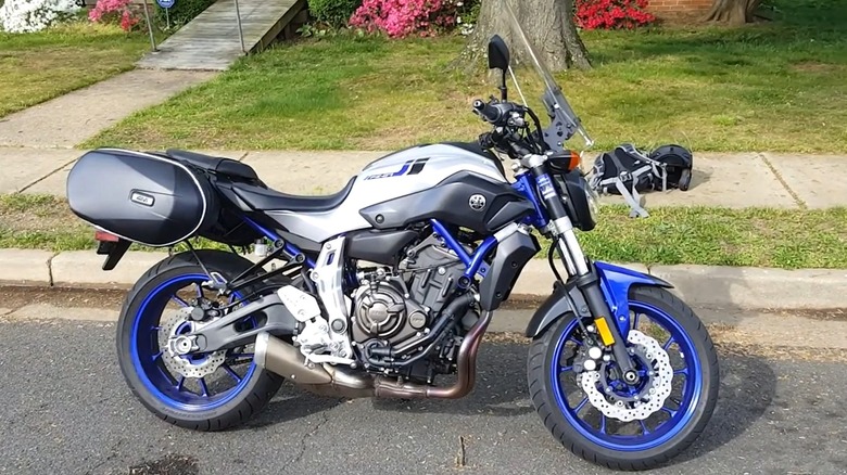 A 2016 Yamaha FZ-07 fitted with saddlebags and a small windshield, is parked by the curb in front of a suburban home on a sunny day.