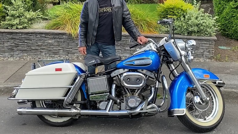 A person in a leather jacket standing beside a vintage blue 1972 Harley Davidson FLH Electra Glide.