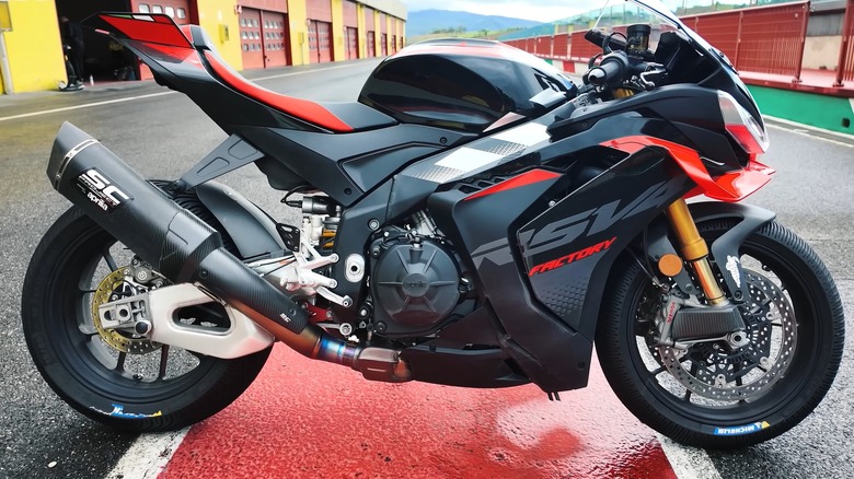 A 2025 Aprilia RSV4 Factory 1100 fitted with an SC Project carbon exhaust sits in a pit lane at what appears to be an Italian circuit.
