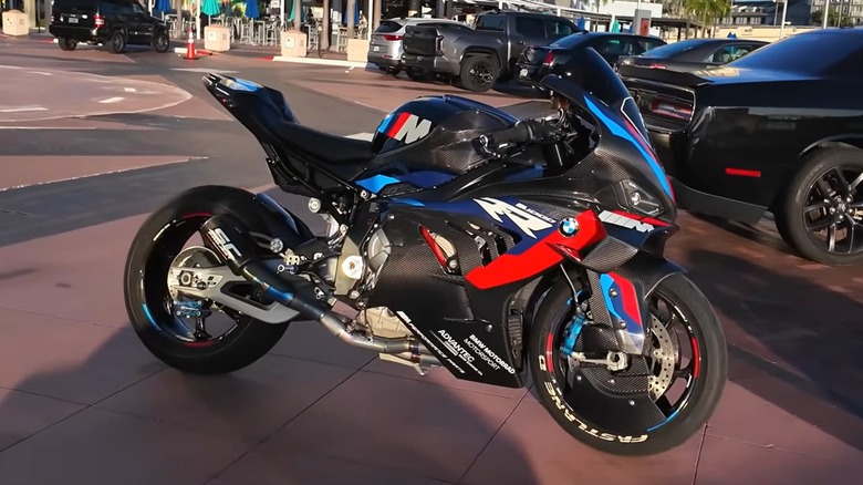 A black BMW M1000RR in M Motorsport livery parked on a waterfront promenade next to a Dodge Challenger