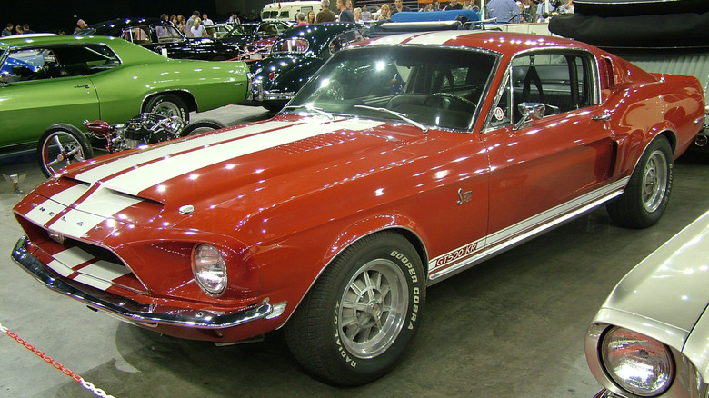 1968 Ford Mustang Shelby GT500 KR parked indoors among classic cars and motorcycles, front-left 3/4 view