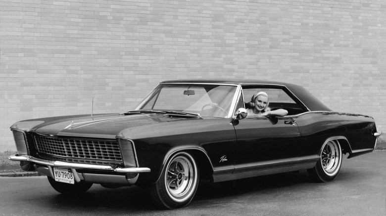 1965 Buick Riviera with woman leaning out of driver's side window, front-left 3/4 view