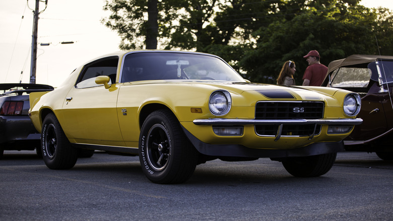 A yellow second generation Chevrolet Camaro with black stripes at a car show, front 3/4 view, sunset