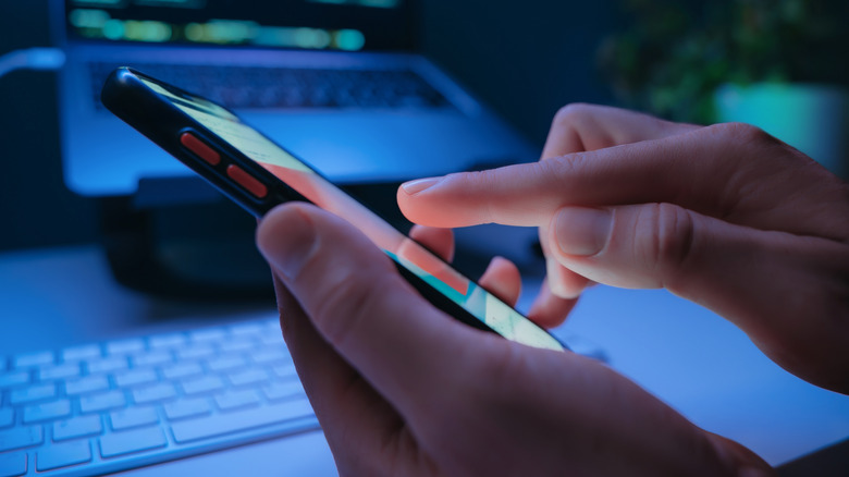 A person scrolling on smartphone with a laptop blurred in the background
