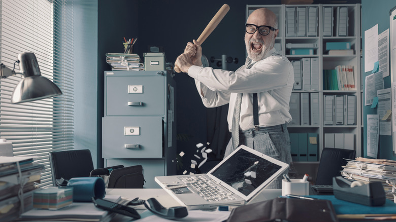 An angry man in an office destroying a laptop with a bat