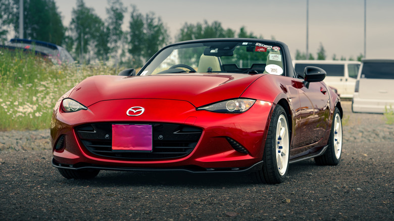 Igora Drive, Red Mazda MX-5 Miata convertible car parked on gravel with white vans and green trees in the background