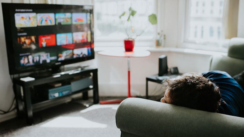 A person in a blue shirt laying on a couch while looking at a smart TV that is showing a selection of films and tv shows to watch.