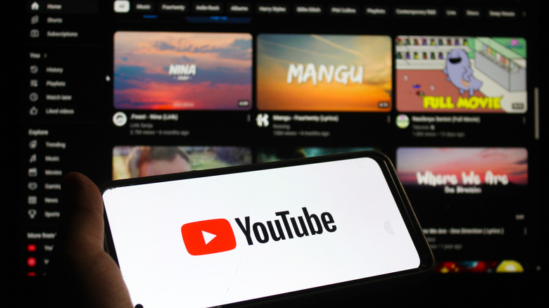 Someone holding a smart phone with the "YouTube" logo on it in front of a YouTube homepage with thumbnails for videos.