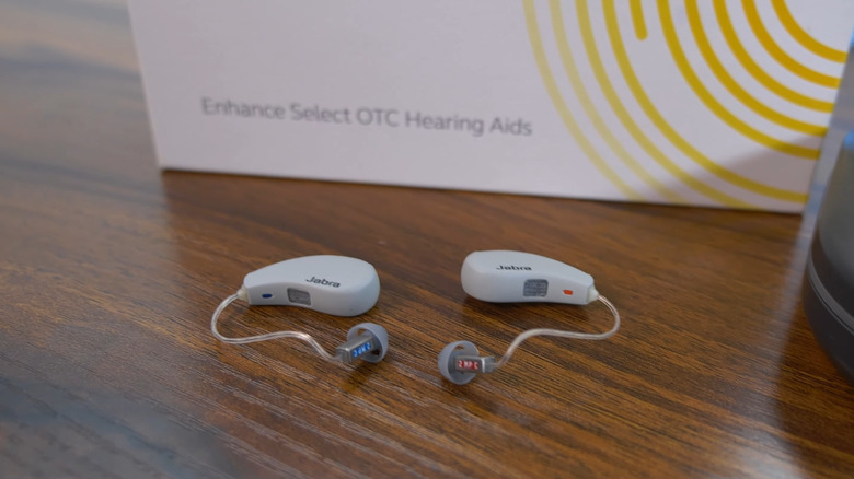 A pair of gray/beige Jabra Enhance Select over-the-counter hearing aids on a wooden table with their box.