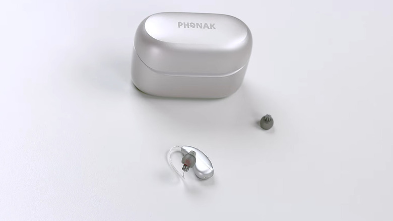 A single gray Phoank hearing aid placed face-up on a white table, with a custom earpiece and silver charging case next to it.