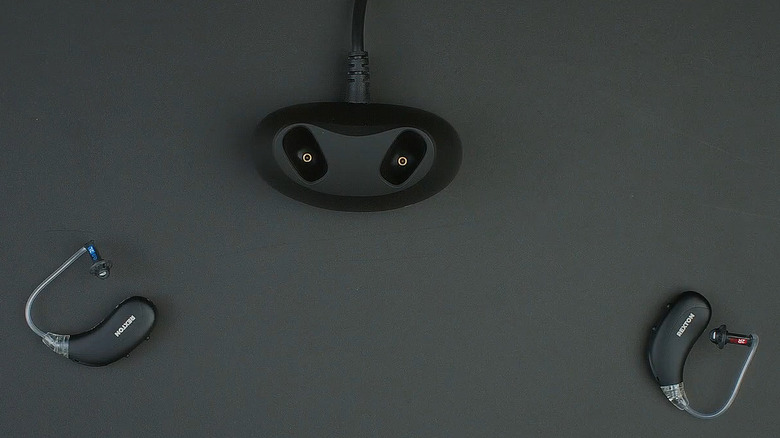 A pair of black and gray Rexton hearing aids laying face-up on a table, with the charging case nearby.