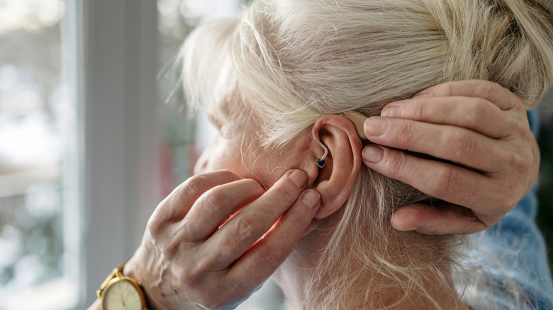 An elderly lady with white hair and her hair in a bun adjusting her BTE (behind-the-ear) hearing aid.