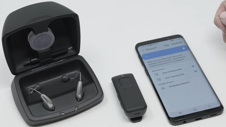 A pair of black Audibel Hearing aids in their case on a white desk, next to a remote and a smartphone.