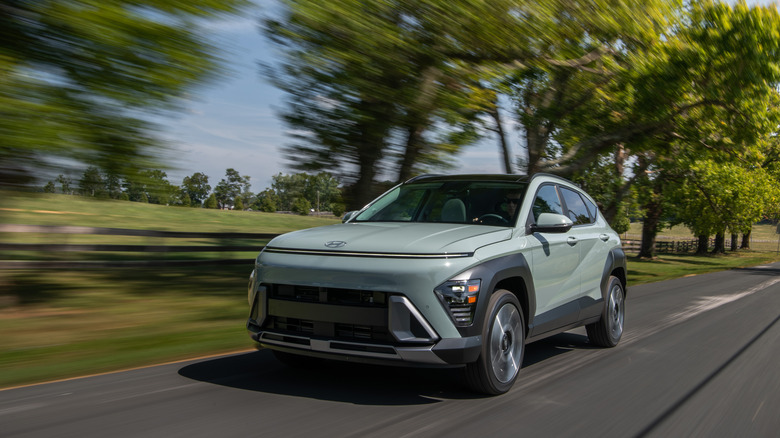 2026 Hyundai Kona driving at speed along country road with trees in background, front-left view