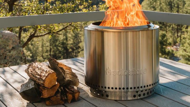 Silver Solo Stove Bonfire with an active fire in the middle, wood on the left of it, on a wooden patio with a forest backdrop