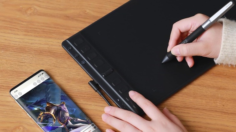 Hand writing on a Huion Inspiroy tablet with a stylus, next to a phone.
