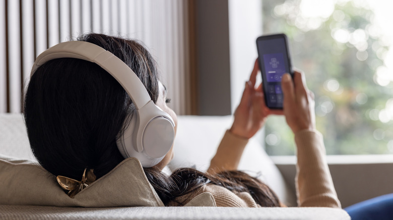 Woman holding her phone and listening to songs on headphones while laying on a couch