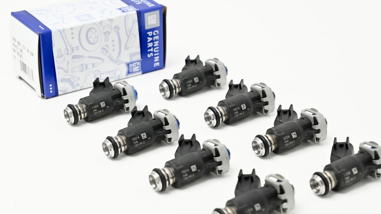 A set of eight Genuine GM fuel injectors arranged in rows on a white surface, with a blue and white GM box in the background